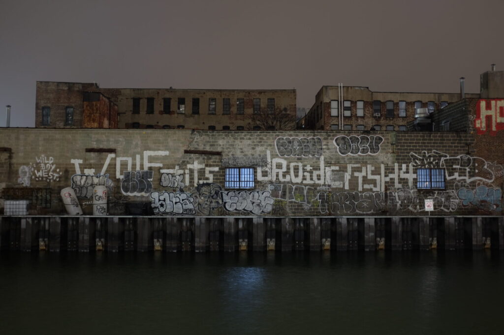 A one story warehouse with crude graffiti tags like "WOLF", "tits", and "dROid" overlooking an industrial canal 