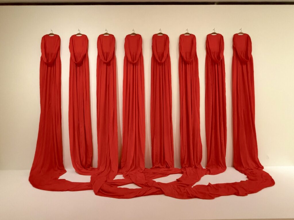 Sixteen orange velvet dresses hanging on metal hangers with their sleeves joined together and long skirts interconnected, draping to the floor to create a wall-like fabric installation